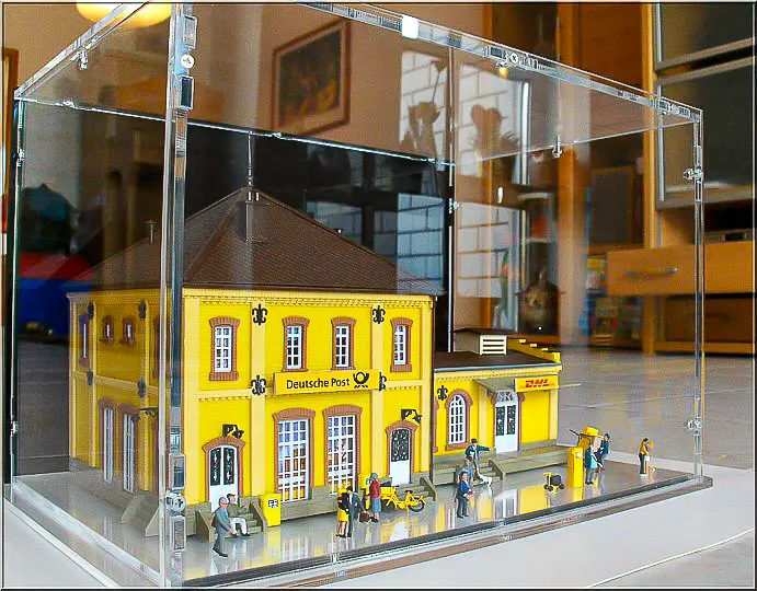 Acryl-Vitrine über Modellbahnhof mit gelber Deutsche-Post-Fassade und kleinen Personenfiguren auf dem Bahnsteig